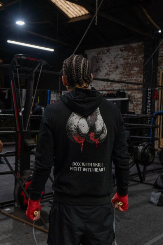 Boxer in a black Prevail hoodie with the boxing gloves graphic and BOX WITH SKILL FIGHT WITH HEART across the back, holding a skipping rope with red hand wraps in the gym